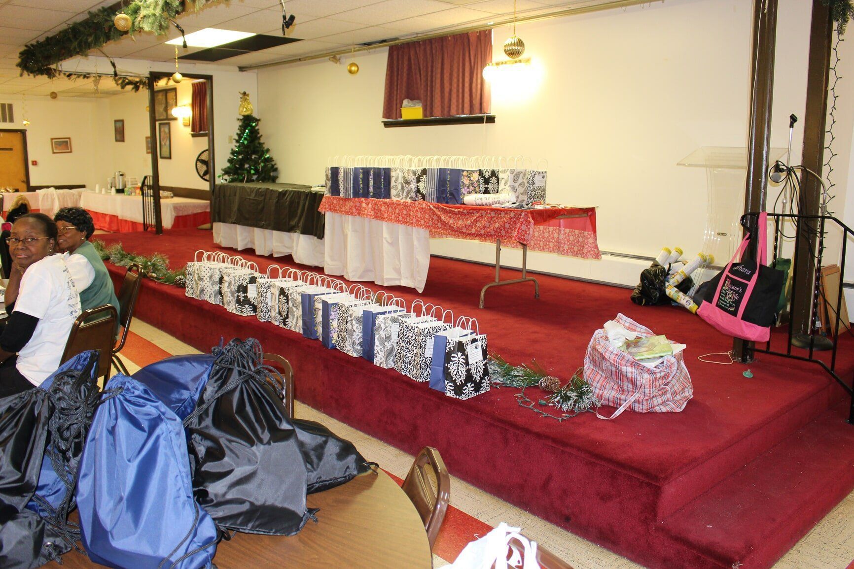 Decorated banquet hall with red-draped tables and a small stage, with gifts and wrapped items in front