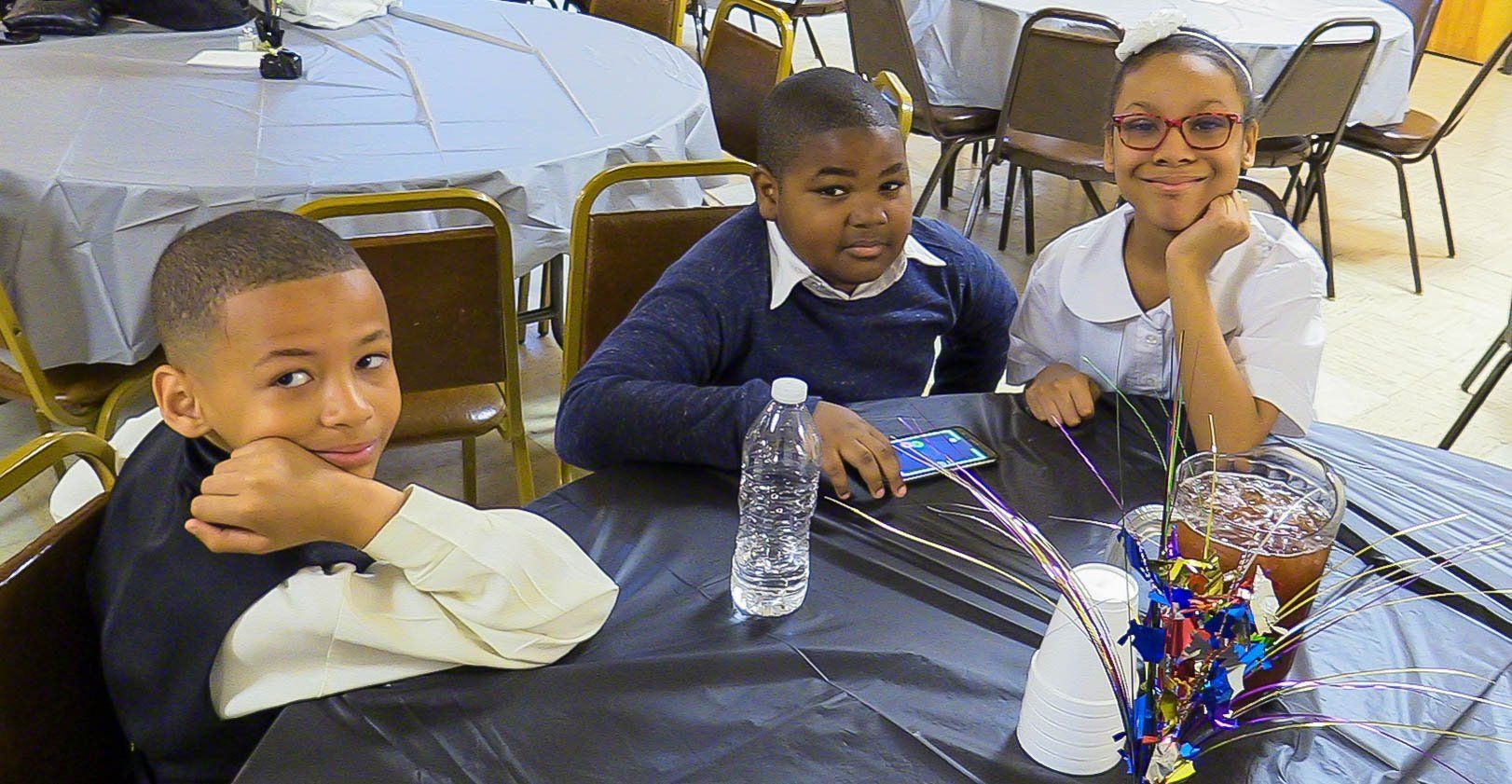 Three children seated at a table with a black tablecloth, smiling beside a water bottle and colorful centerpiece.
