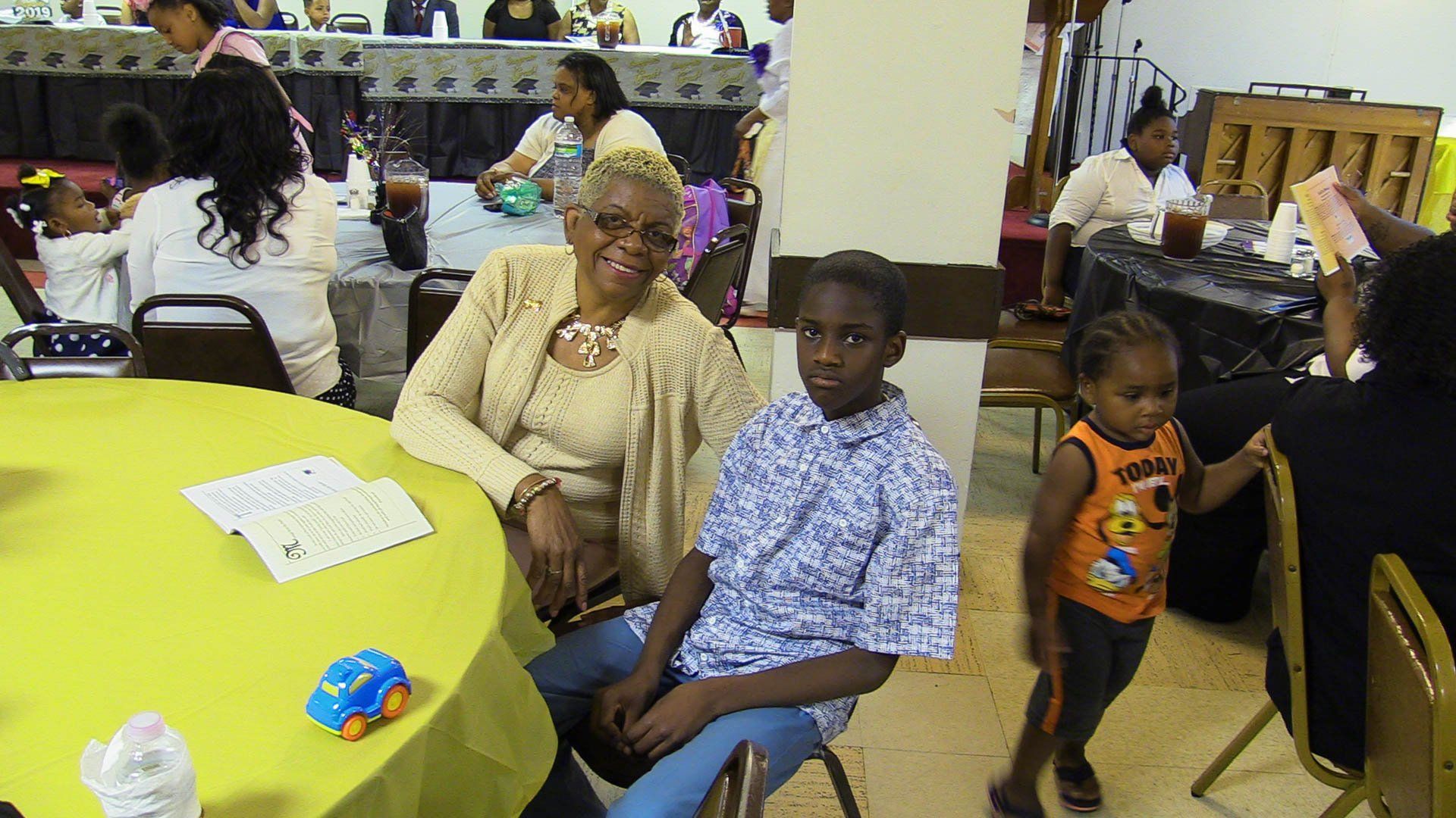 People seated at a round yellow table in a busy hall, with children standing nearby and vending machines in back.