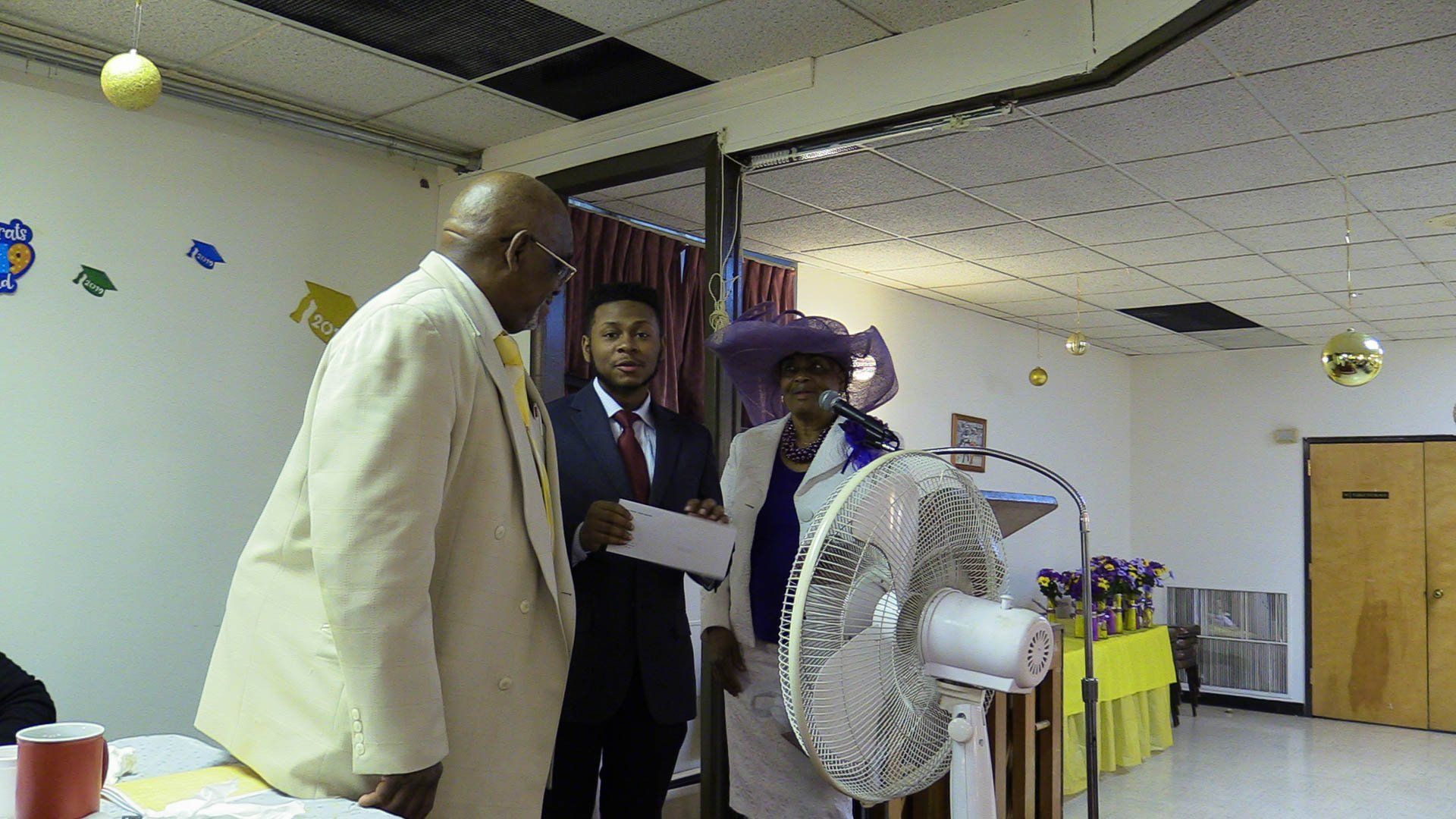 People standing at an indoor event beside a large white fan, with a yellow table and wall decorations in the background
