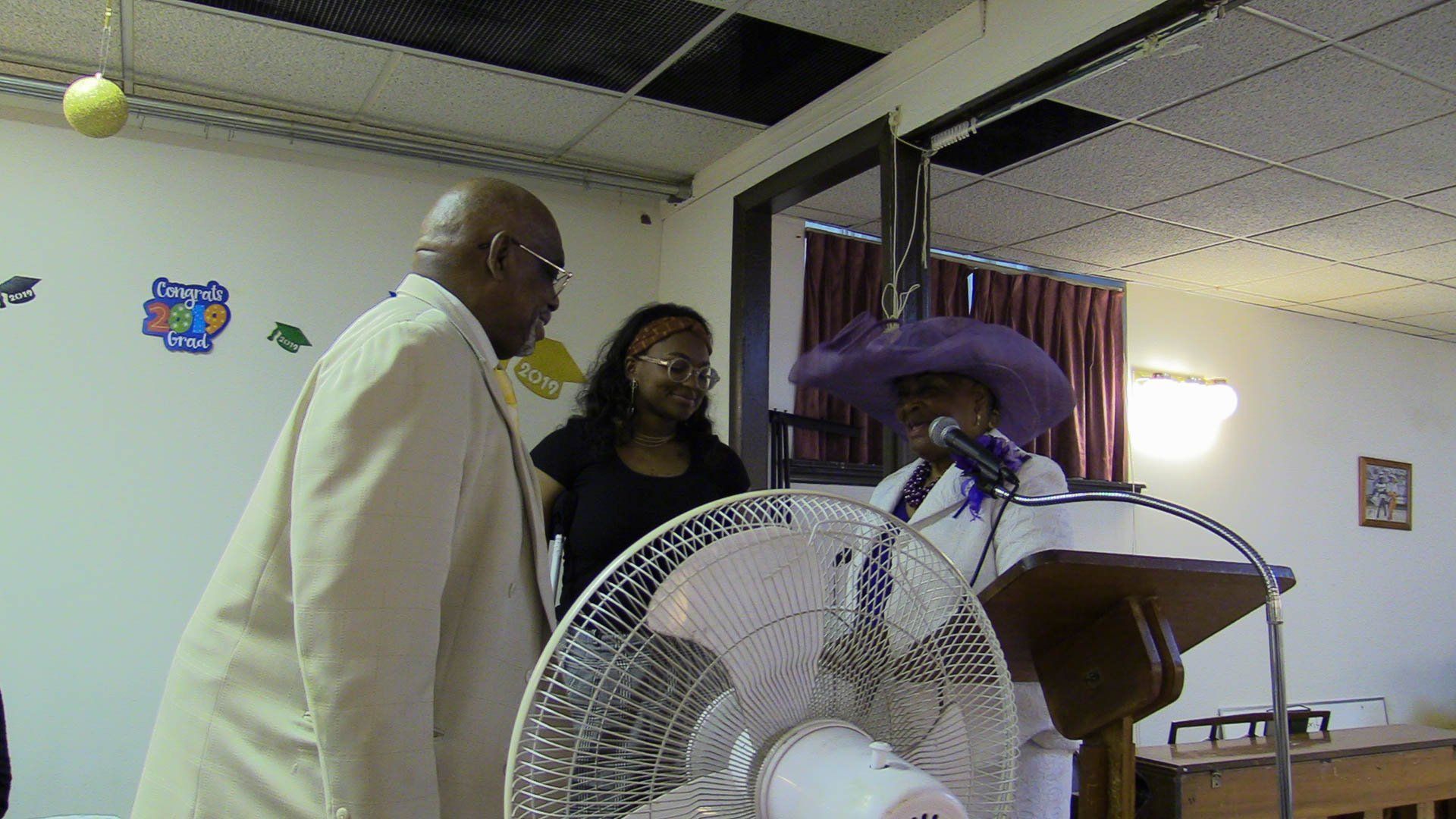 Two people stand beside a lectern and fan in a small indoor room during a presentation.
