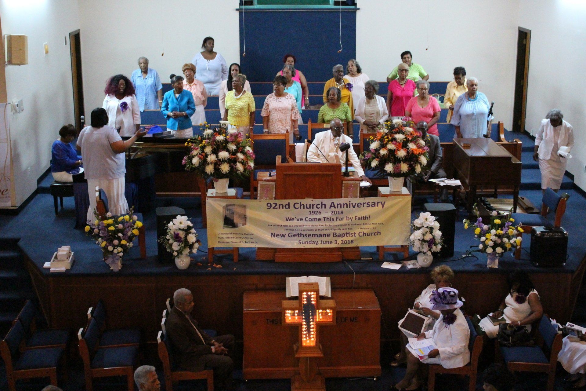 Choir standing on a church stage behind flower arrangements and a banner during a service