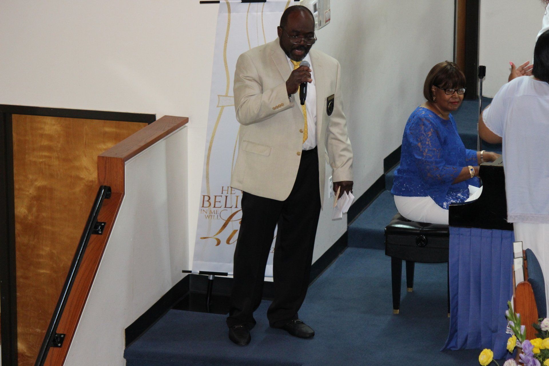 Man speaking into a microphone at a podium beside a piano in a church-like hall
