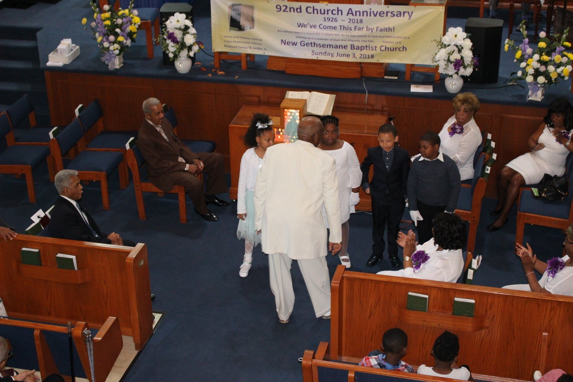 People in a church aisle during a ceremony, with pews and floral decorations visible.