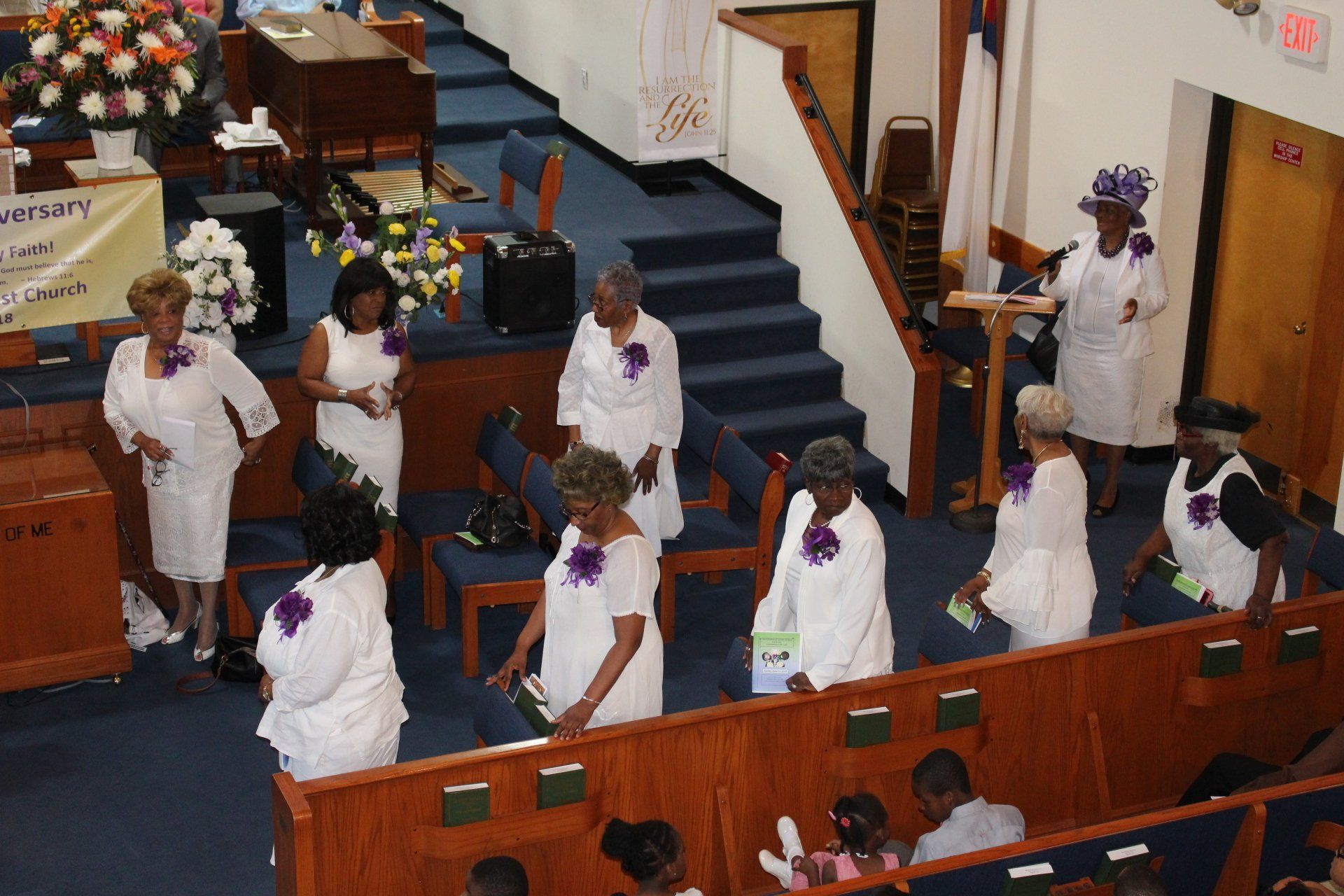 People in white robes stand in a church aisle near the pulpit and stairs.