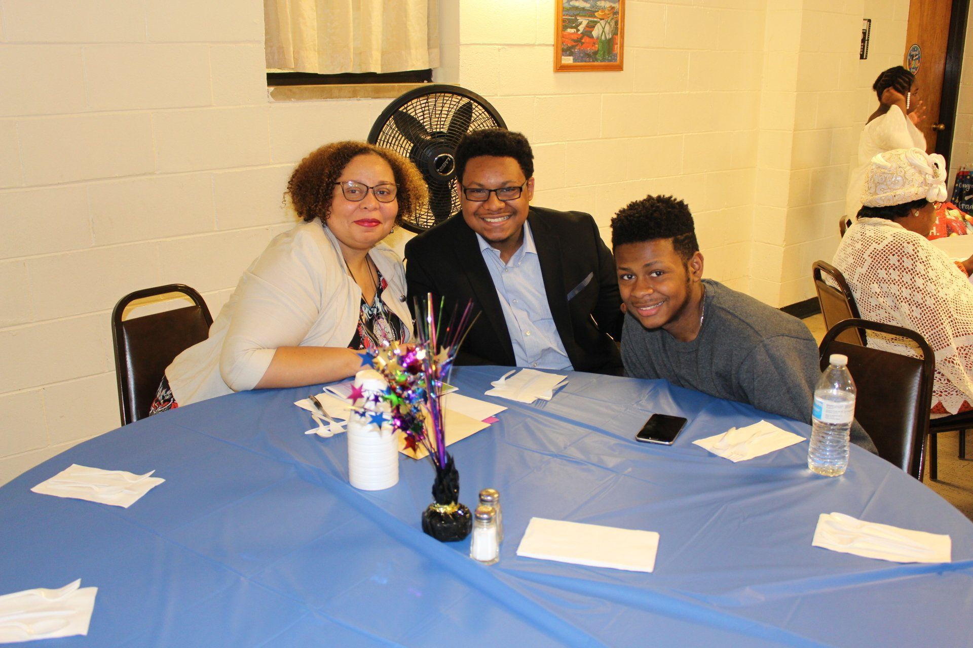 Three people smiling at a blue-clothed banquet table with flowers and papers in a hall.