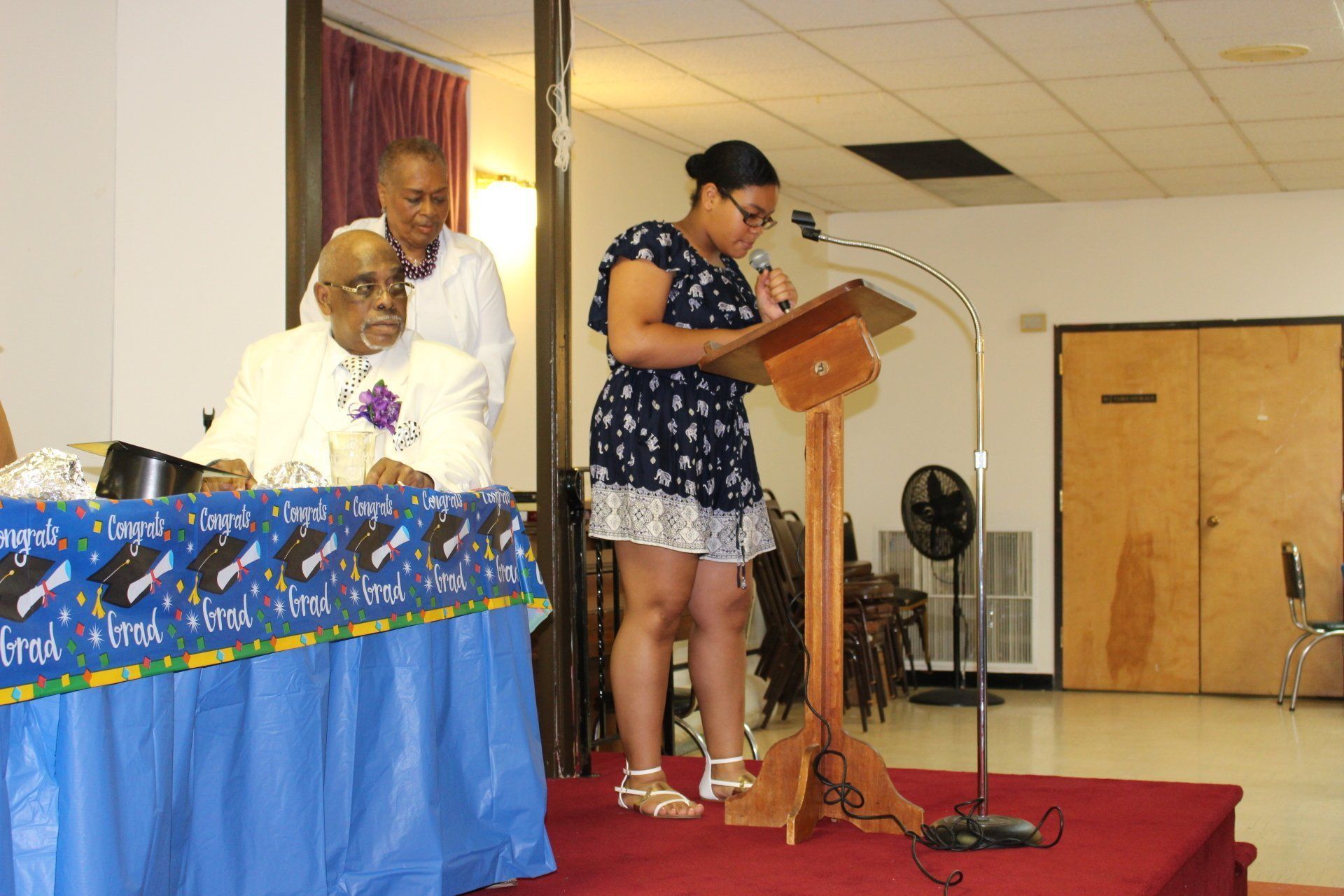 Woman speaking at a podium on a small stage while two people stand nearby; table with blue cloth in foreground
