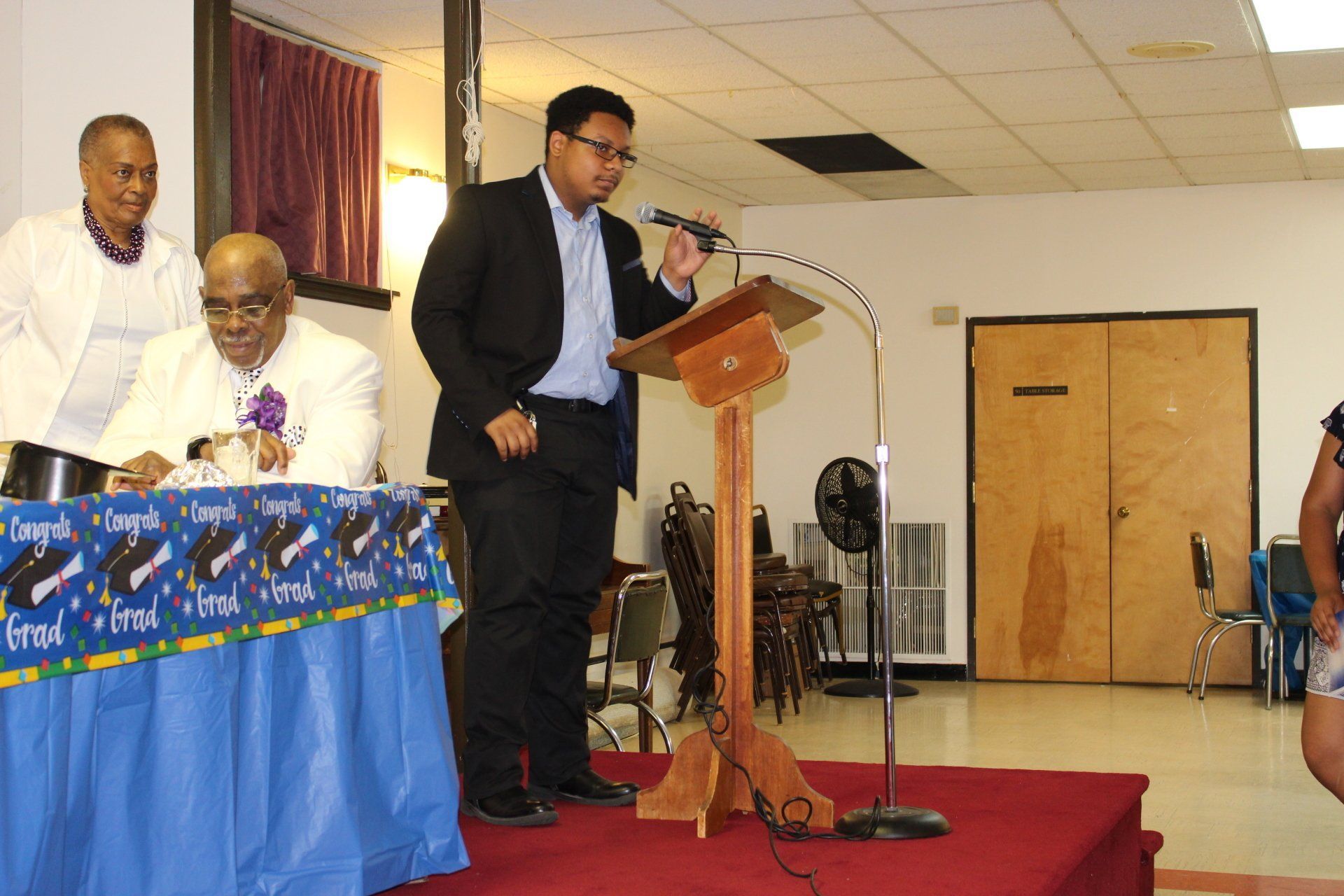 Man speaking at a podium in a community hall, with seated panelists and a blue tablecloth nearby.