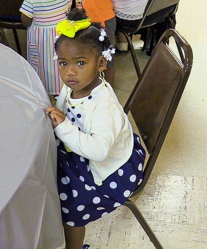 Child in white cardigan and navy polka-dot dress seated at a table, looking at the camera.