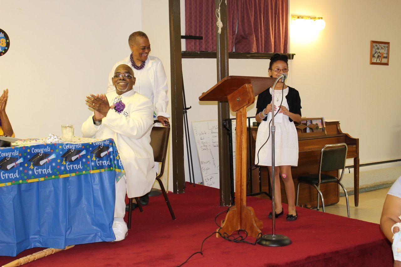 Speaker at podium in church hall beside seated guests and a blue-draped table