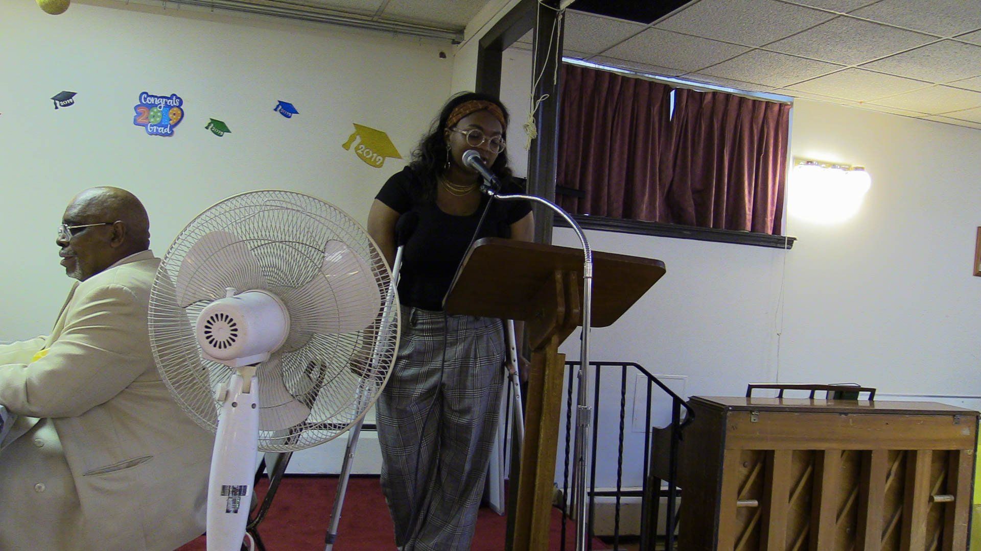 Woman speaking at a church podium beside a fan, with another person seated nearby and a piano in the background
