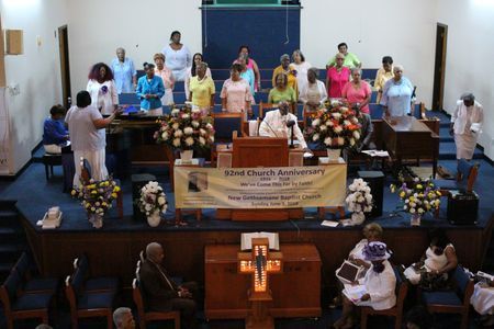 Choir members standing on a church altar during a service, with floral arrangements and a cross in front.