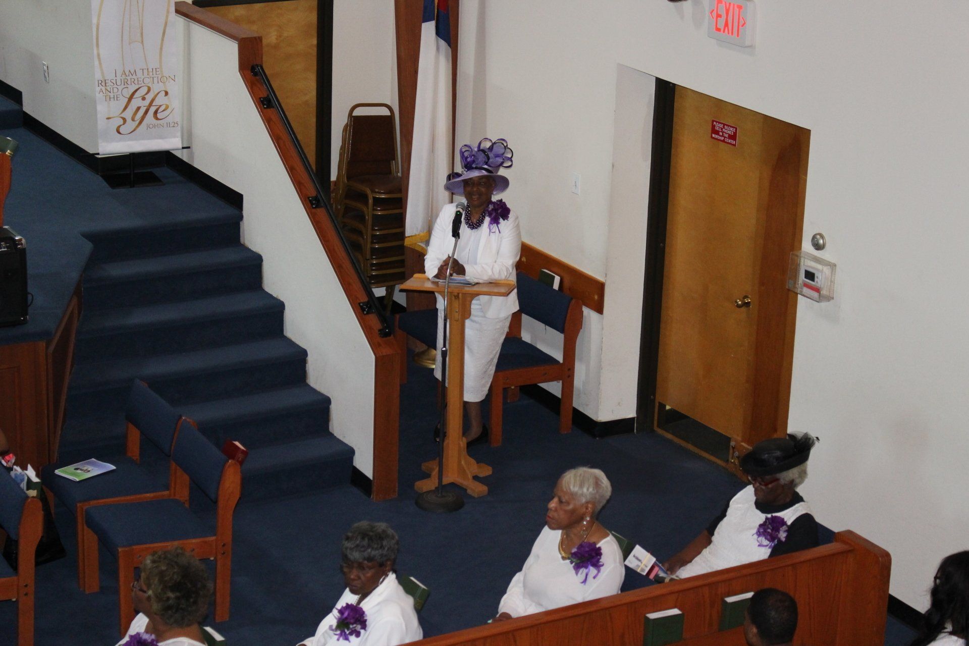 People seated in a church pew near a staircase; a person stands at a pulpit with purple flowers.
