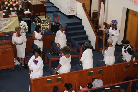 People in white outfits stand in a church aisle during a ceremony.
