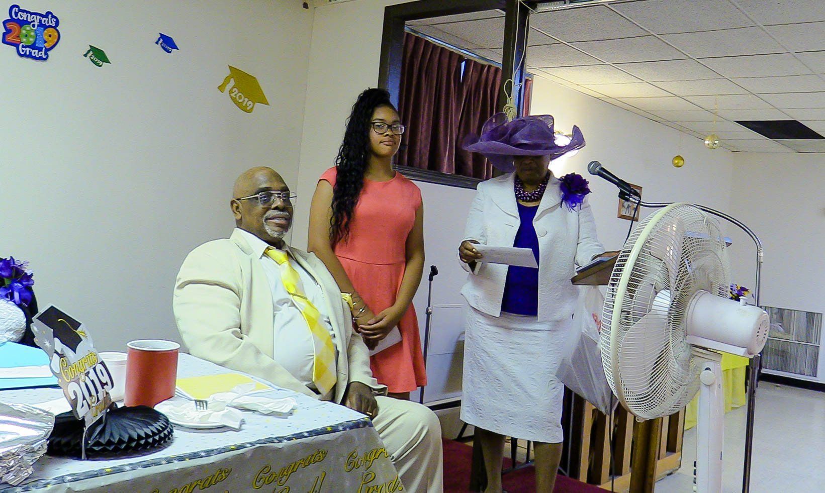 Indoor gathering with three people in formal attire beside a decorated table and fan