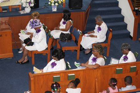 People seated in a church pew, wearing white outfits with purple accents, facing the front.