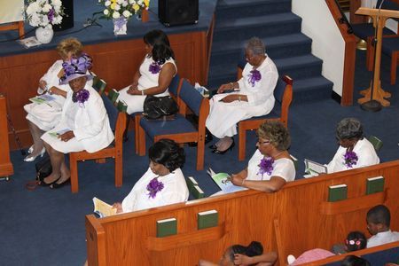 People seated in church pews wearing white outfits with purple accents, reading programs during a service