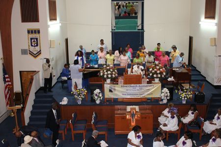Church hall with people seated and standing around a stage filled with colorful flowers and banners.