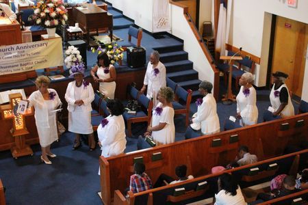 People in white robes gather in a church aisle during a service, with pews and a pulpit nearby.