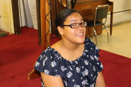 Woman with glasses smiling in a navy patterned top, seated indoors on a red carpet.