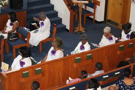People seated in a church pew and on a stage, many wearing white robes with purple sashes.