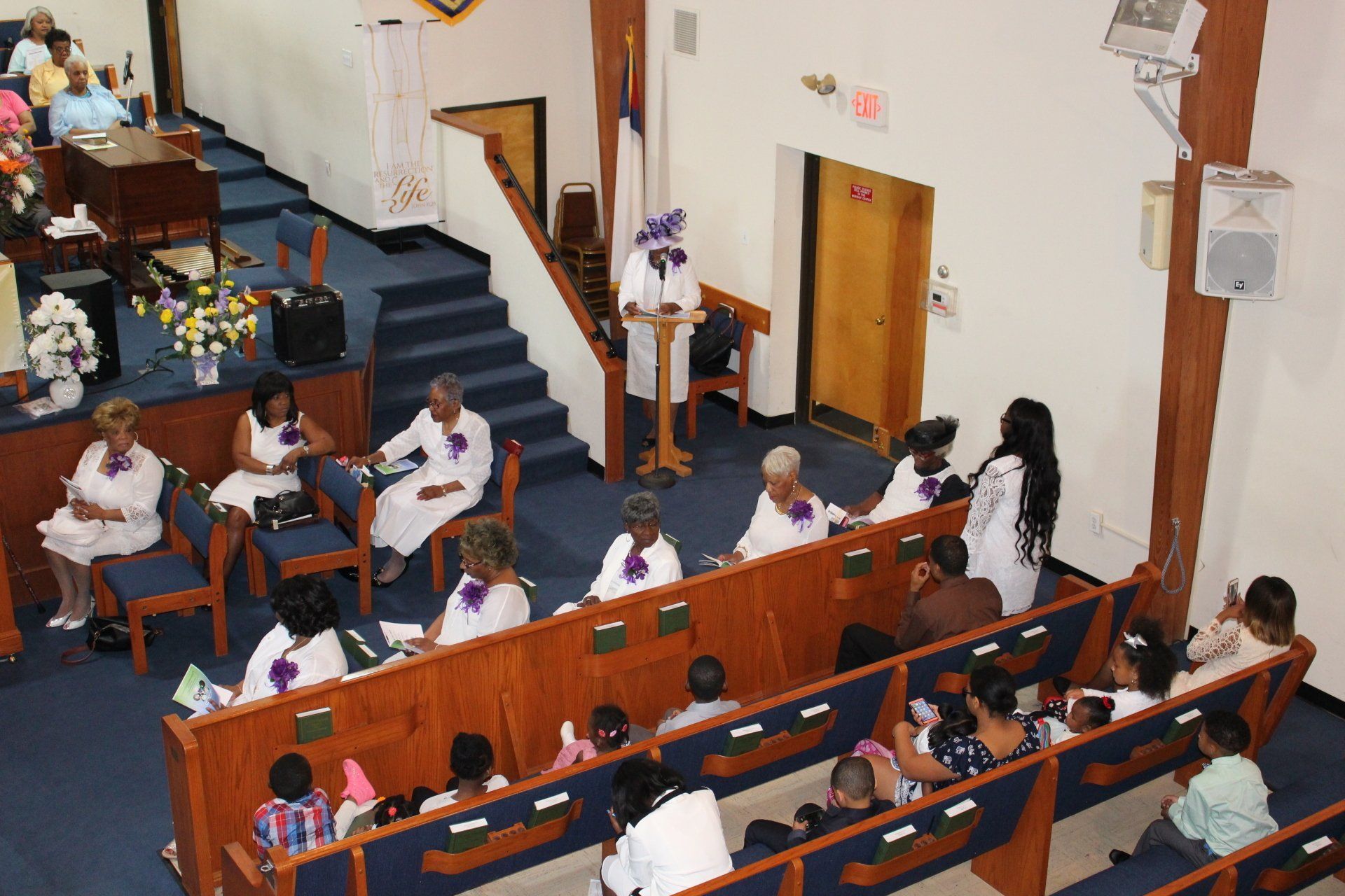 People seated in a church-like hall, viewing a white-clad speaker near the front pews.