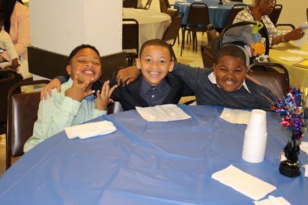 Three smiling children seated at a table with a blue tablecloth in a banquet hall.
