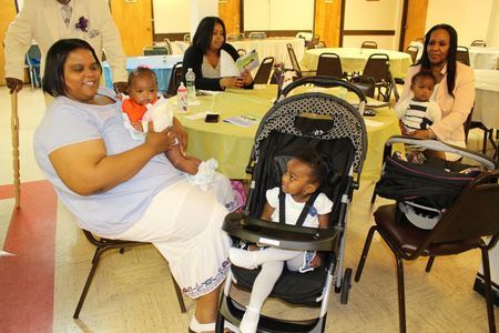 People seated at a table in a banquet hall, with a woman holding a baby and a child in a stroller in front.