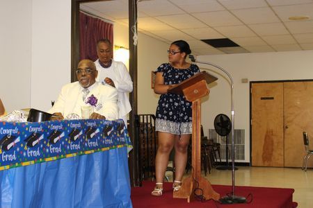 Two speakers at a podium on a small stage beside a table with a blue cloth in a hall