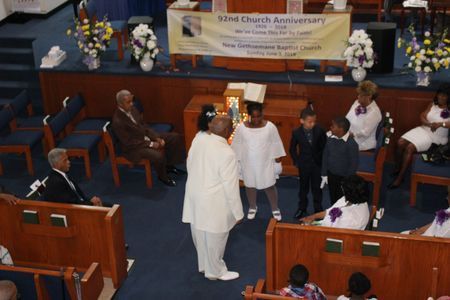 Church service with people standing around a central figure in white, altar flowers, and a “7th Church Anniversary” banner