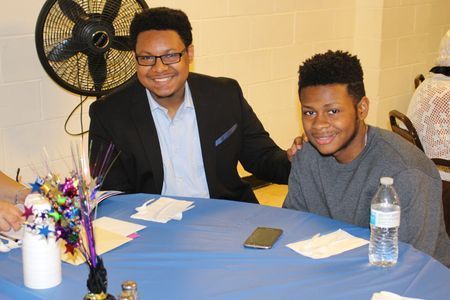 Two people seated at a table with a blue cloth, smiling at an event with a fan and water bottle nearby
