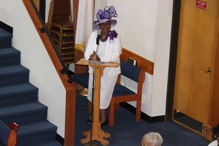 Person standing at a podium in a hallway, wearing a white outfit and purple hat.