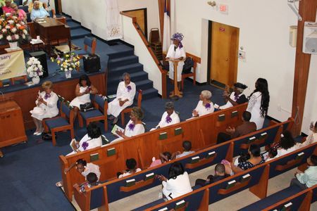 People seated in a church pews during a service, with a woman standing at the front near the aisle.