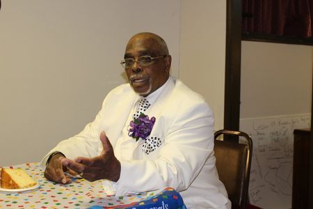 Man in white suit seated at a table with a slice of cake, gesturing while speaking