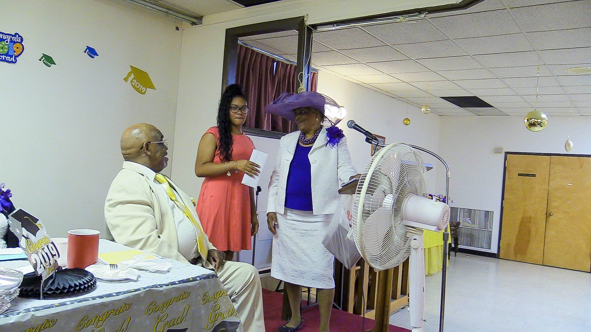 Indoor event with a person speaking into a microphone beside a large fan and seated attendees