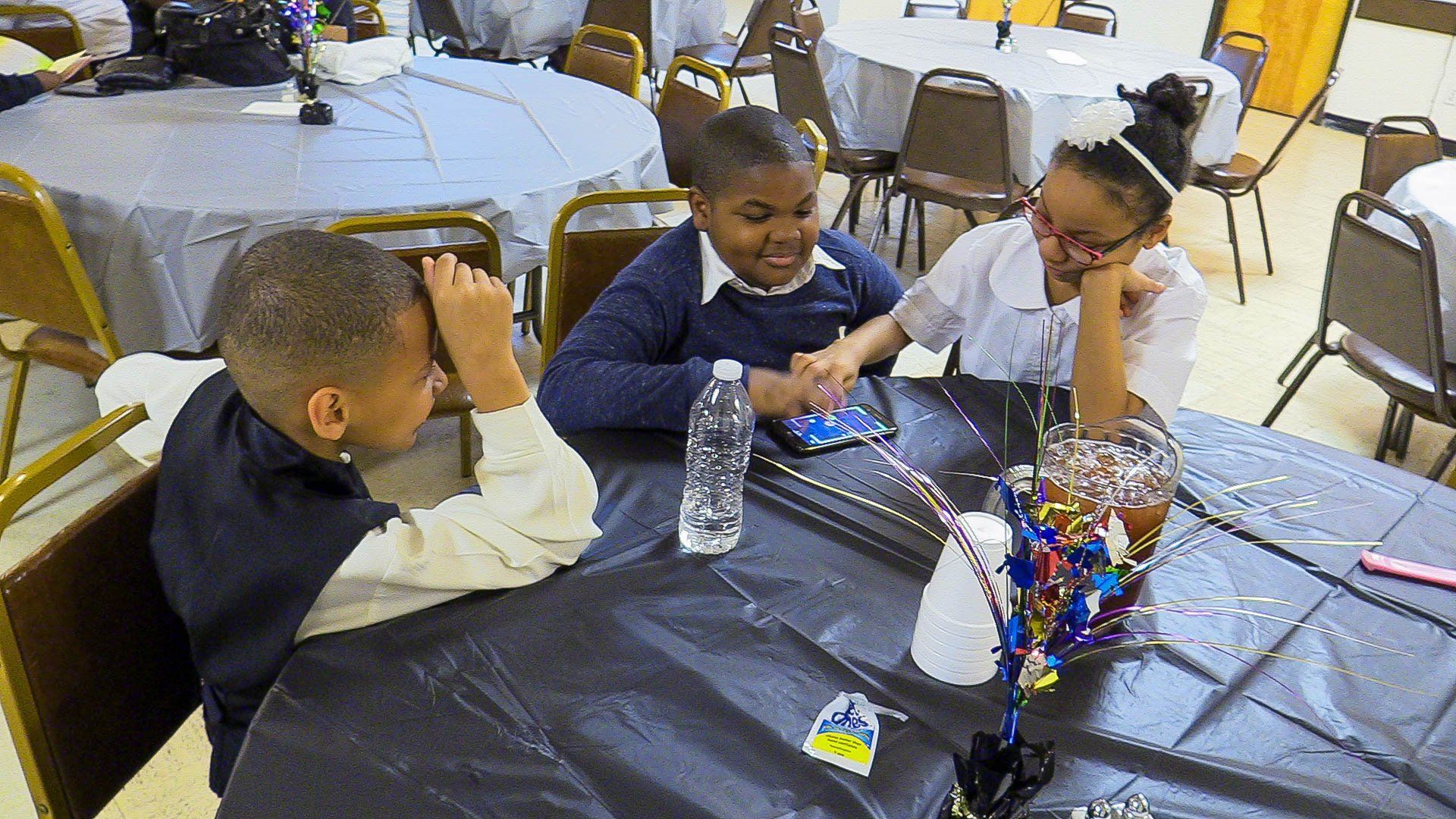 Three children talking at a party table with a gray tablecloth and colorful centerpiece