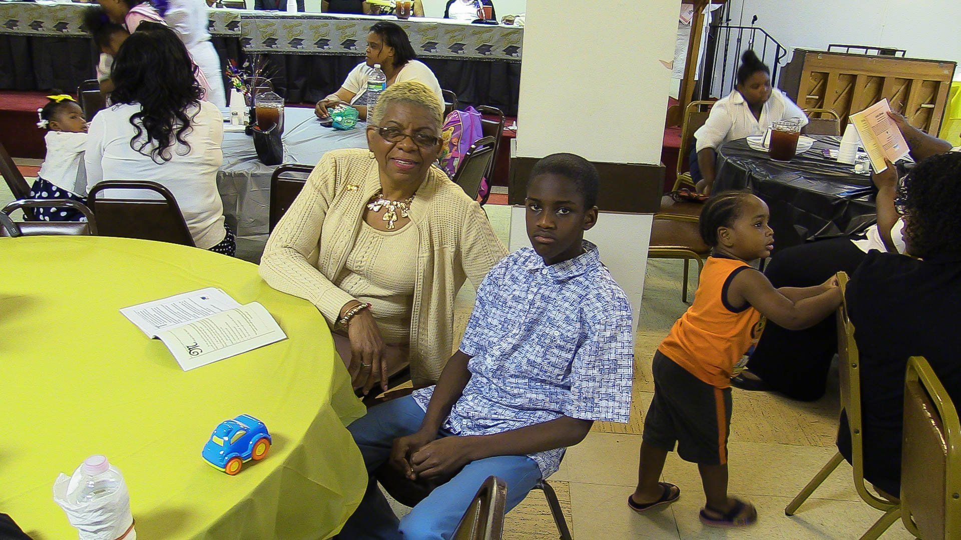 People seated at a yellow table in a community hall, chatting and eating together.