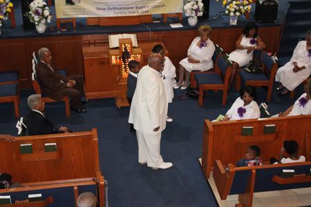 A church service with a speaker in white standing in the aisle while seated congregants watch nearby.