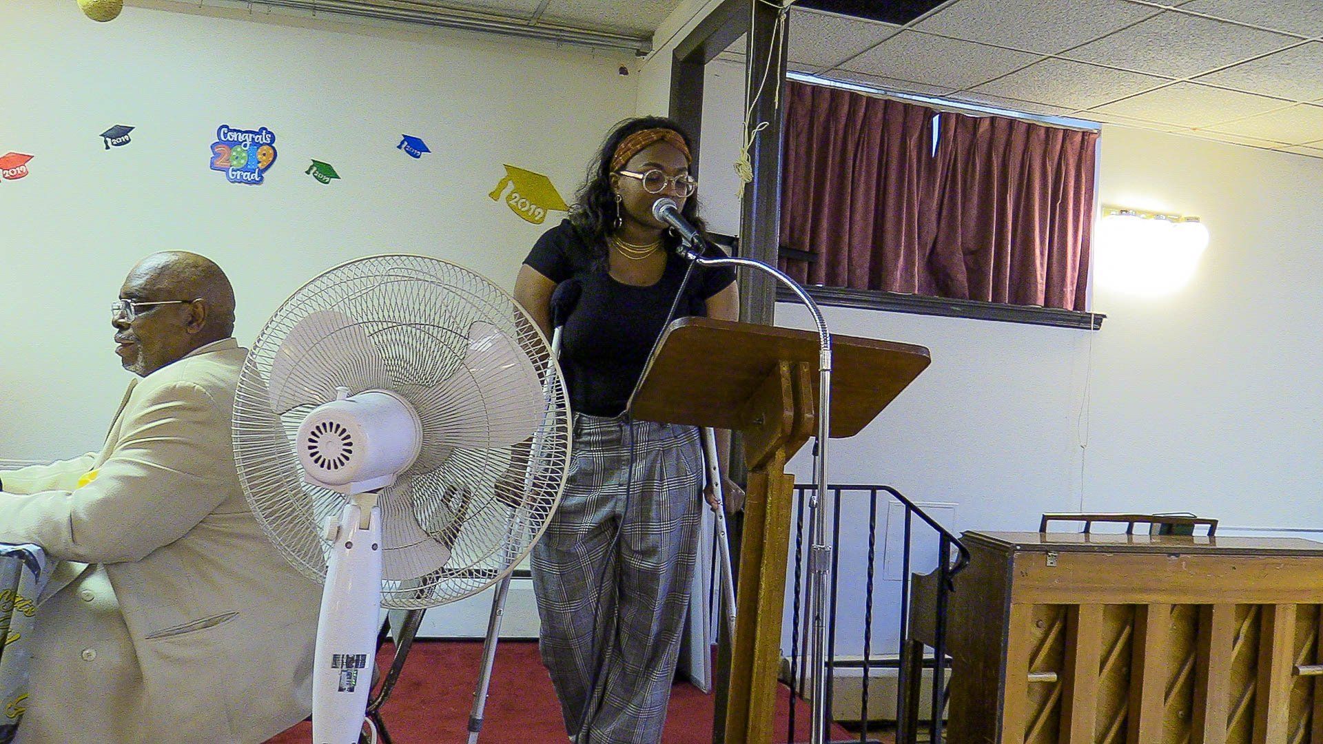 Woman speaking at a podium in a church hall while a man sits nearby.