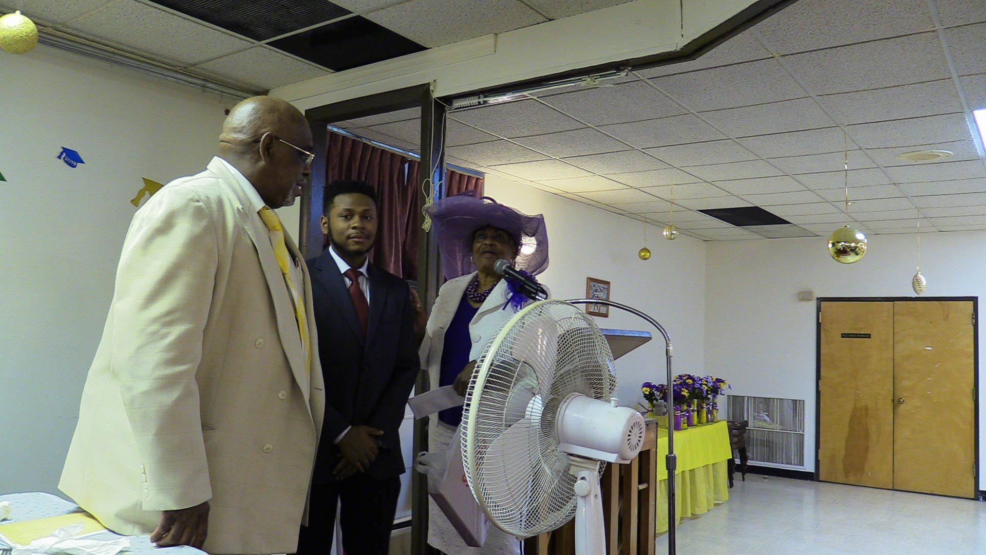Three people stand in a hall near a table, with a large standing fan in the foreground.