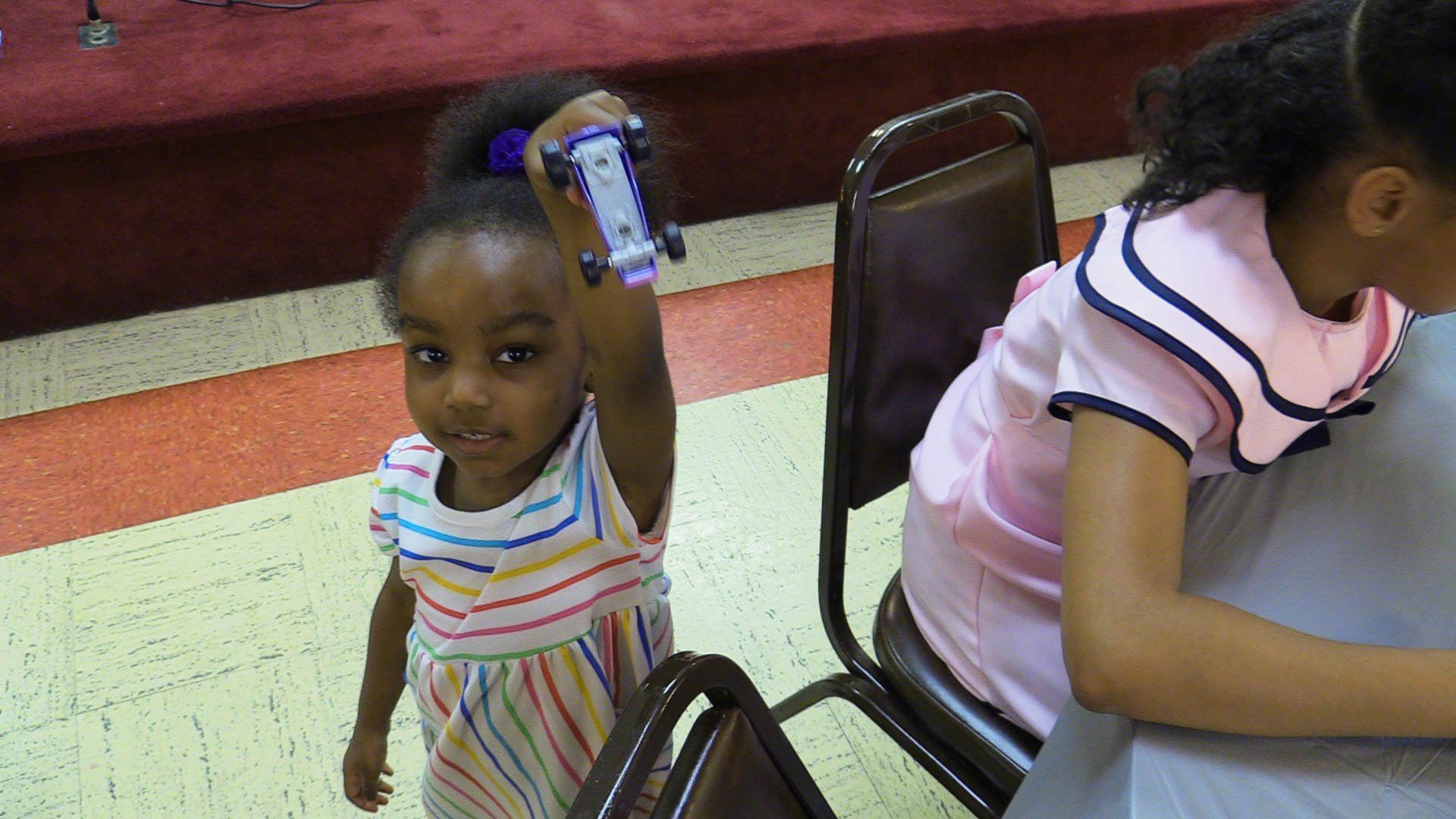 Two children seated at a table, with one child holding a bottle and looking at the camera.