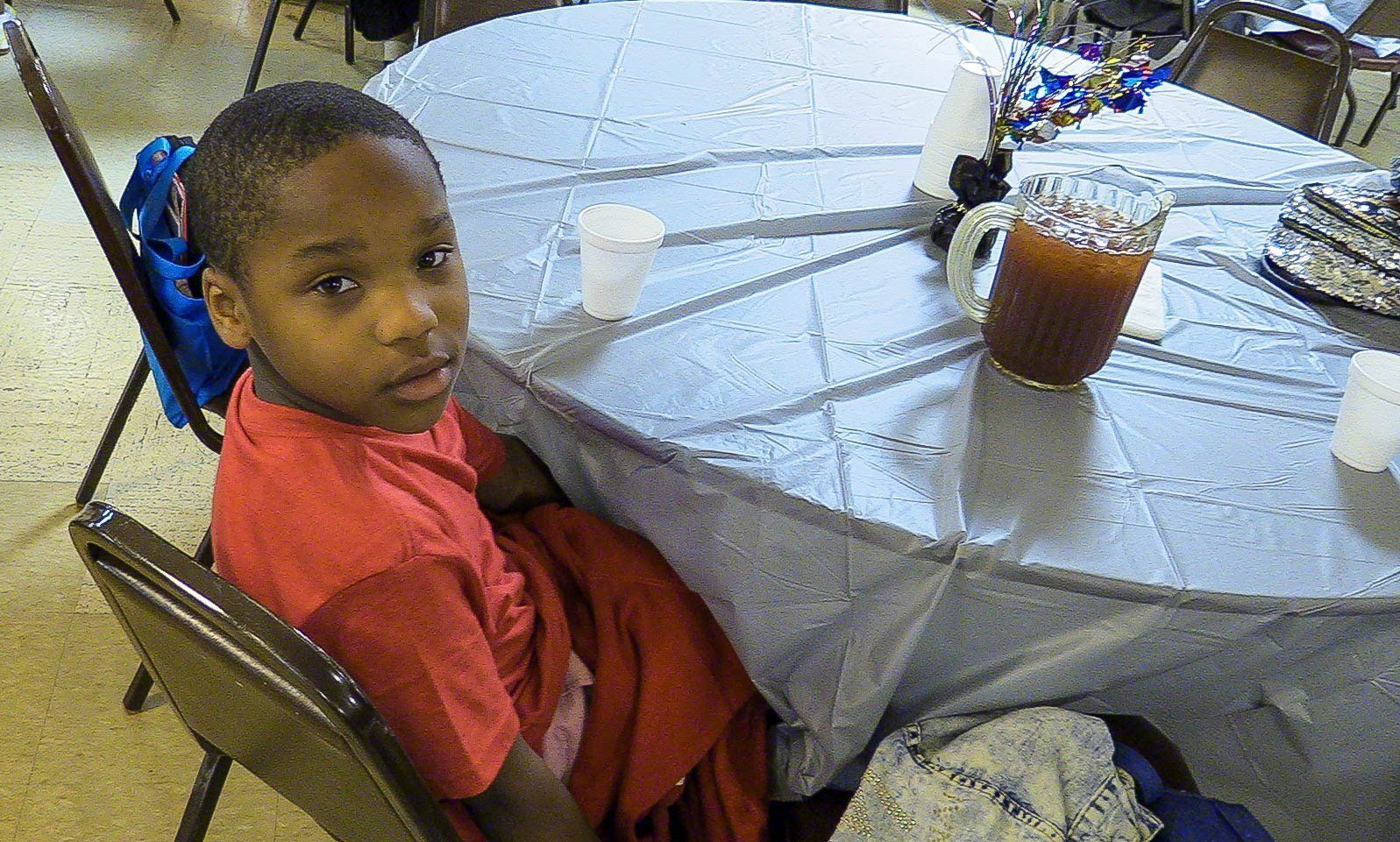 Child in red shirt sitting at a round outdoor table with a cup of iced drink and a black bag.