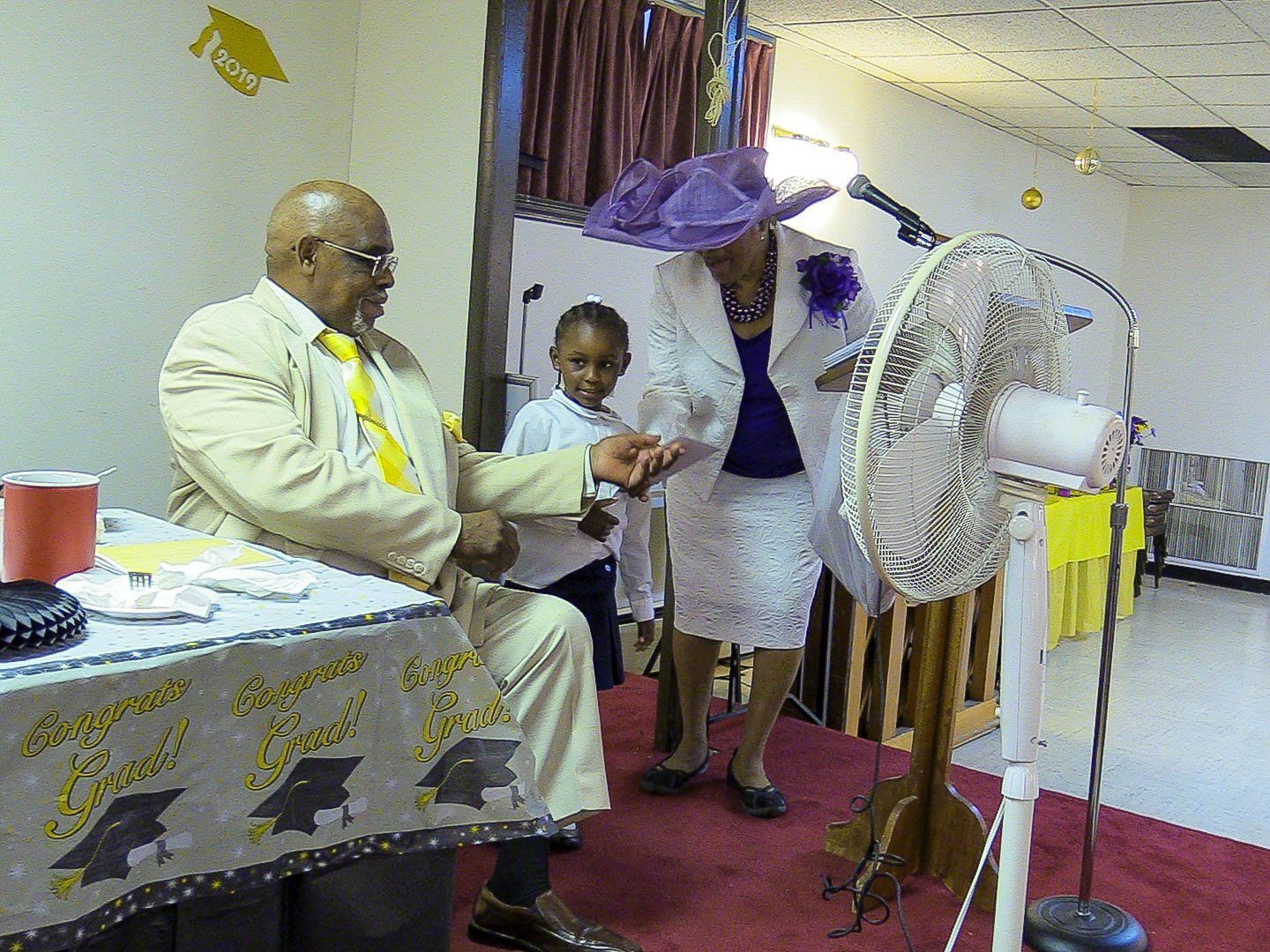 People in a decorated room, with a standing fan and table nearby, during a formal gathering