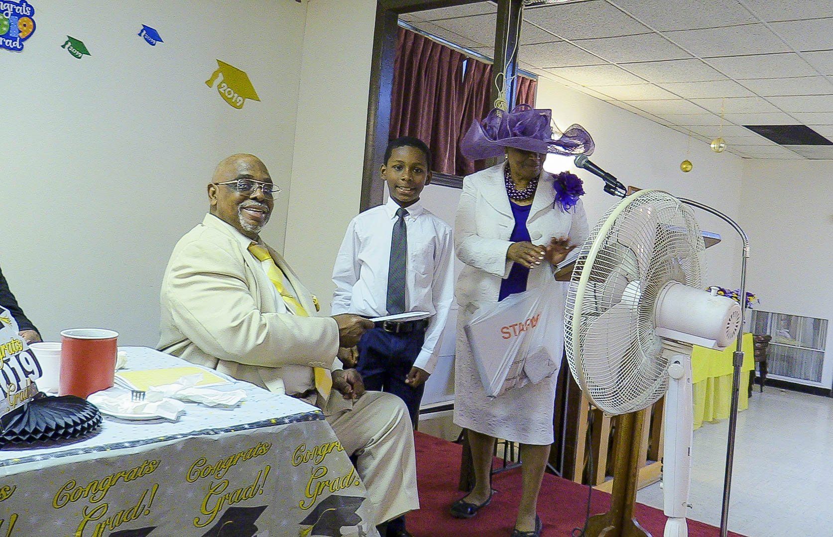 Hospital ceremony with two men seated and one standing beside a decorative fan and purple flowers