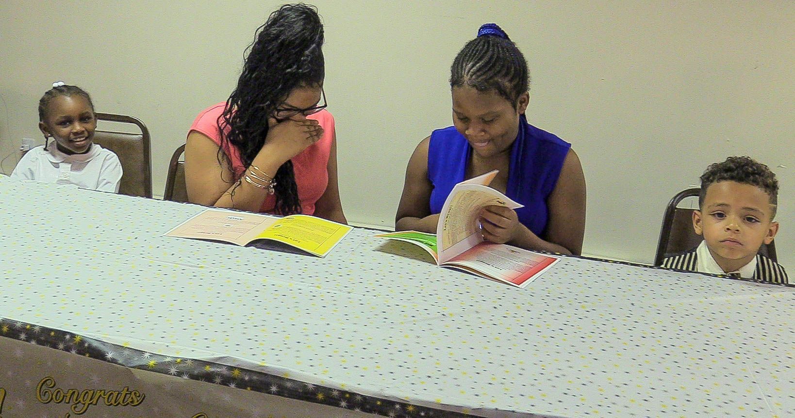 Four children sit at a long table, reading and writing with colorful papers in front of them.