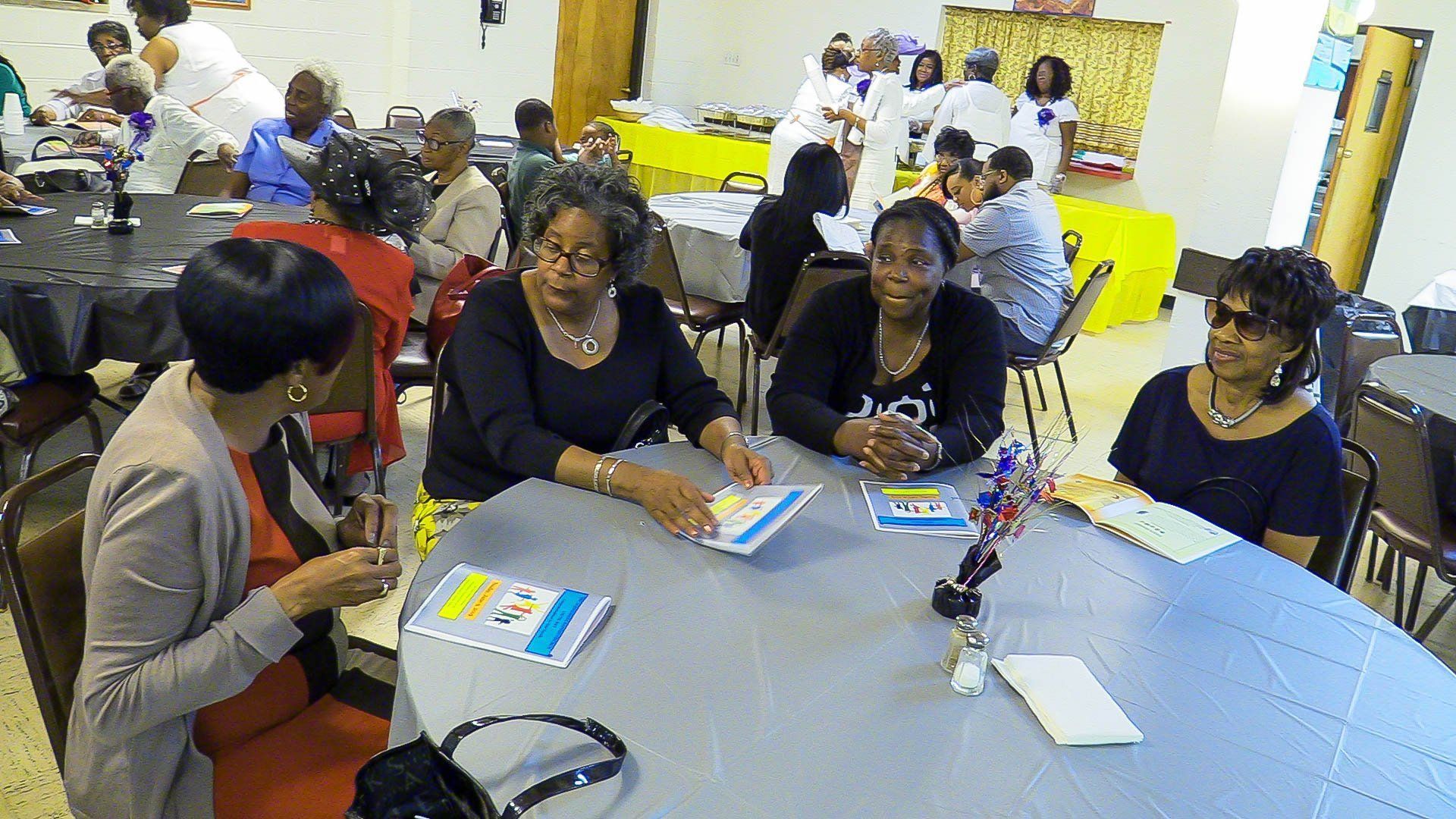 People seated at round tables in a bright meeting room, writing and talking during a group workshop.