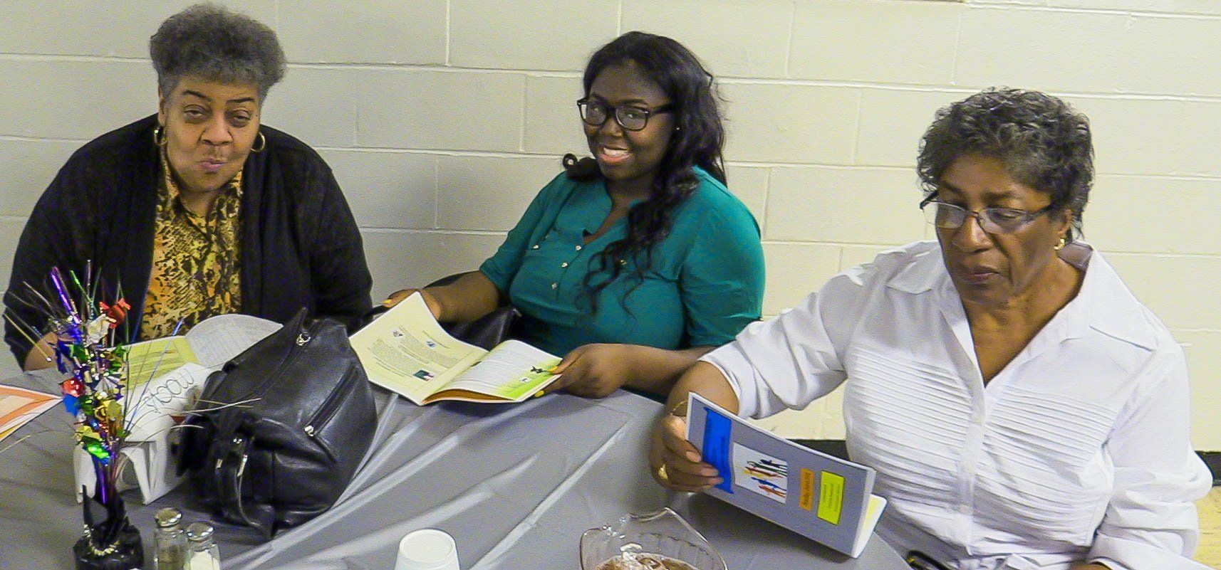 Three people sit at a table with papers and supplies, appearing to work on a project.