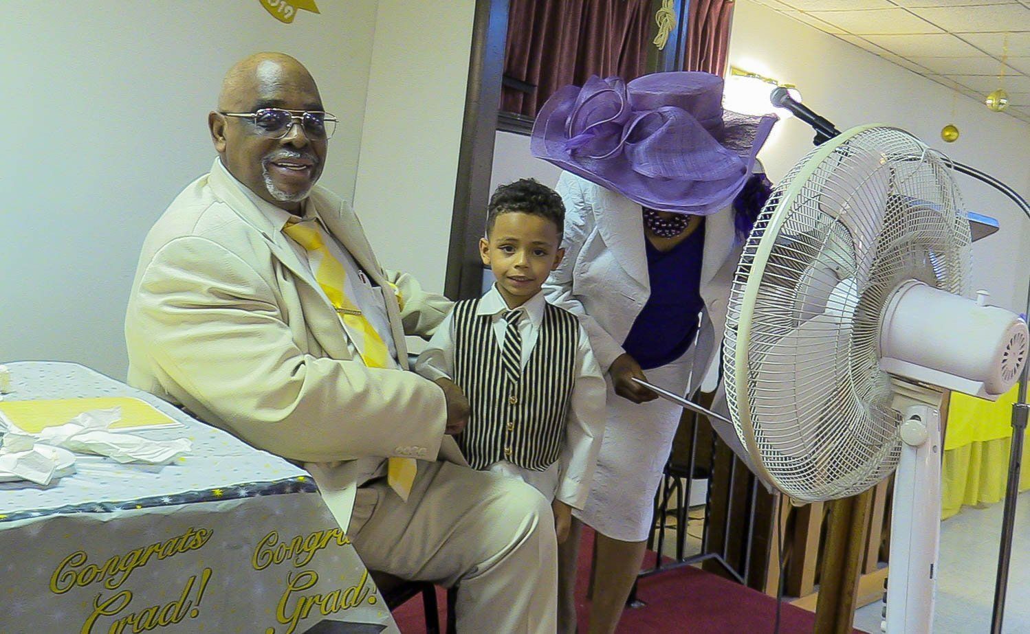Man in a beige suit seated beside a young child in a striped shirt, indoors near a fan and purple decoration.