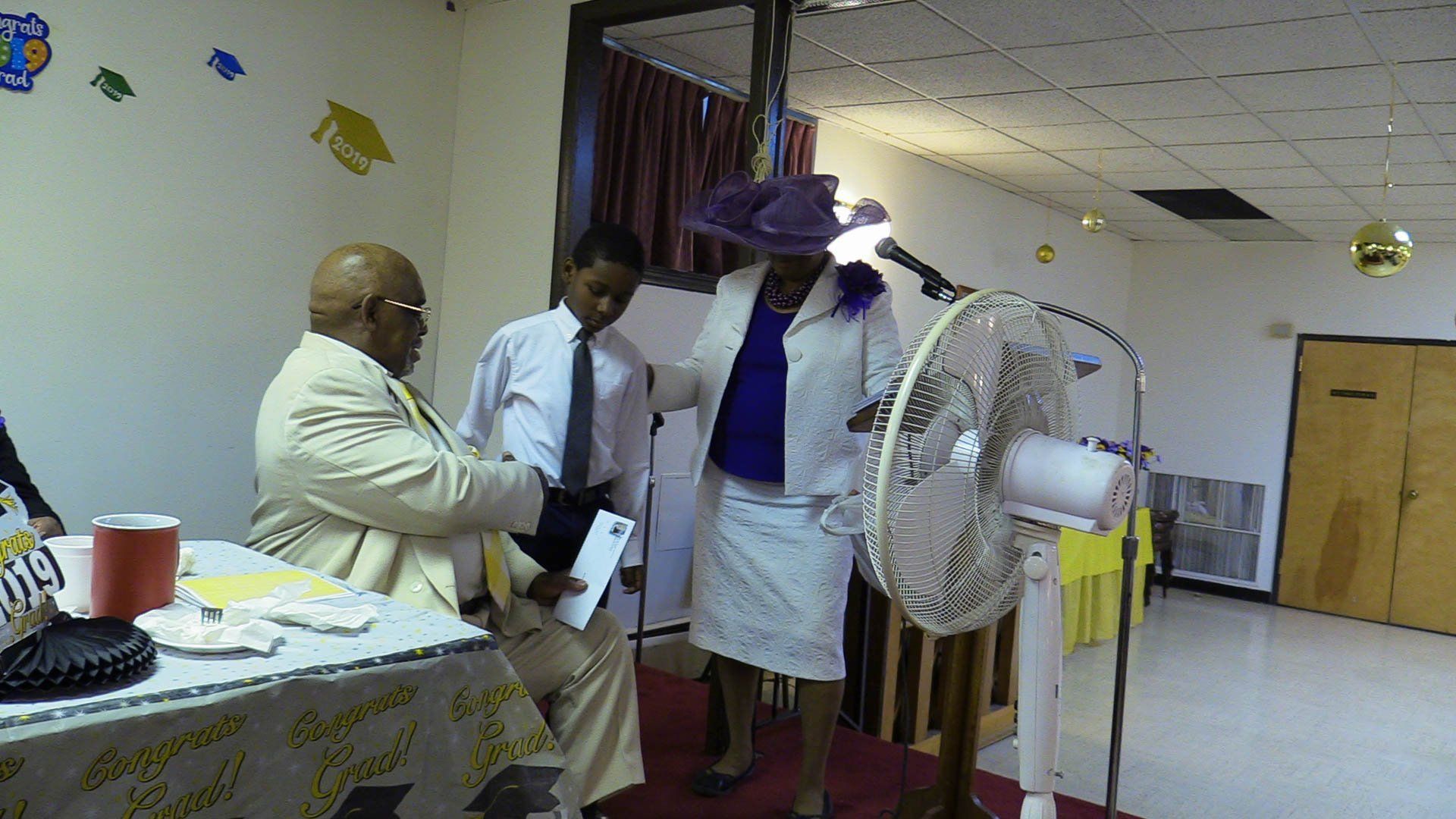 Several people in a room with a fan, table, and posters on the wall