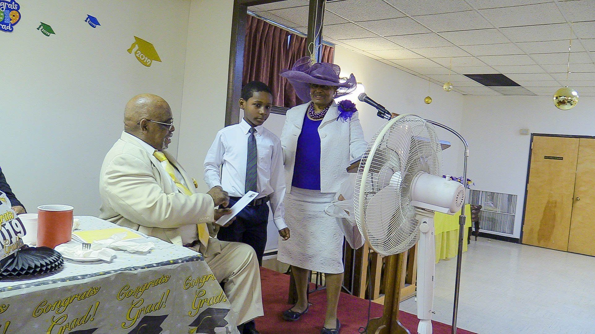 Indoor ceremony with a seated speaker and two people standing near a decorated podium and fan